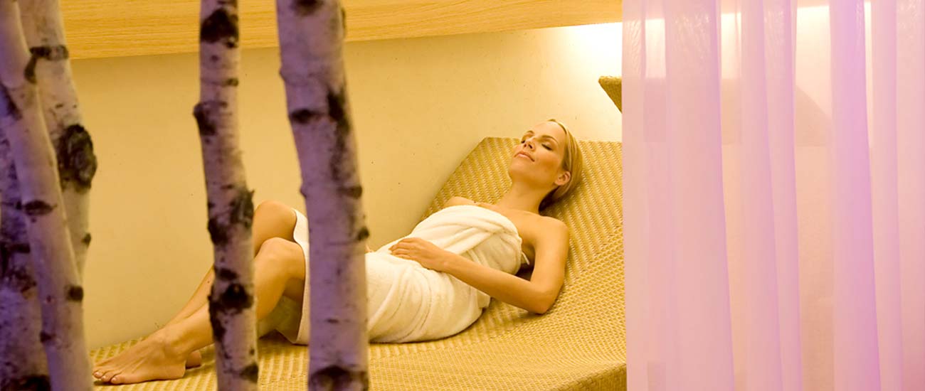 Eine blonde Dame entspannt auf einer Ratanliege im Wellnessbereich der Genusshotels. Im rechten Vordergrund ist eine fliederfarbenes Tuch zu erkennen.