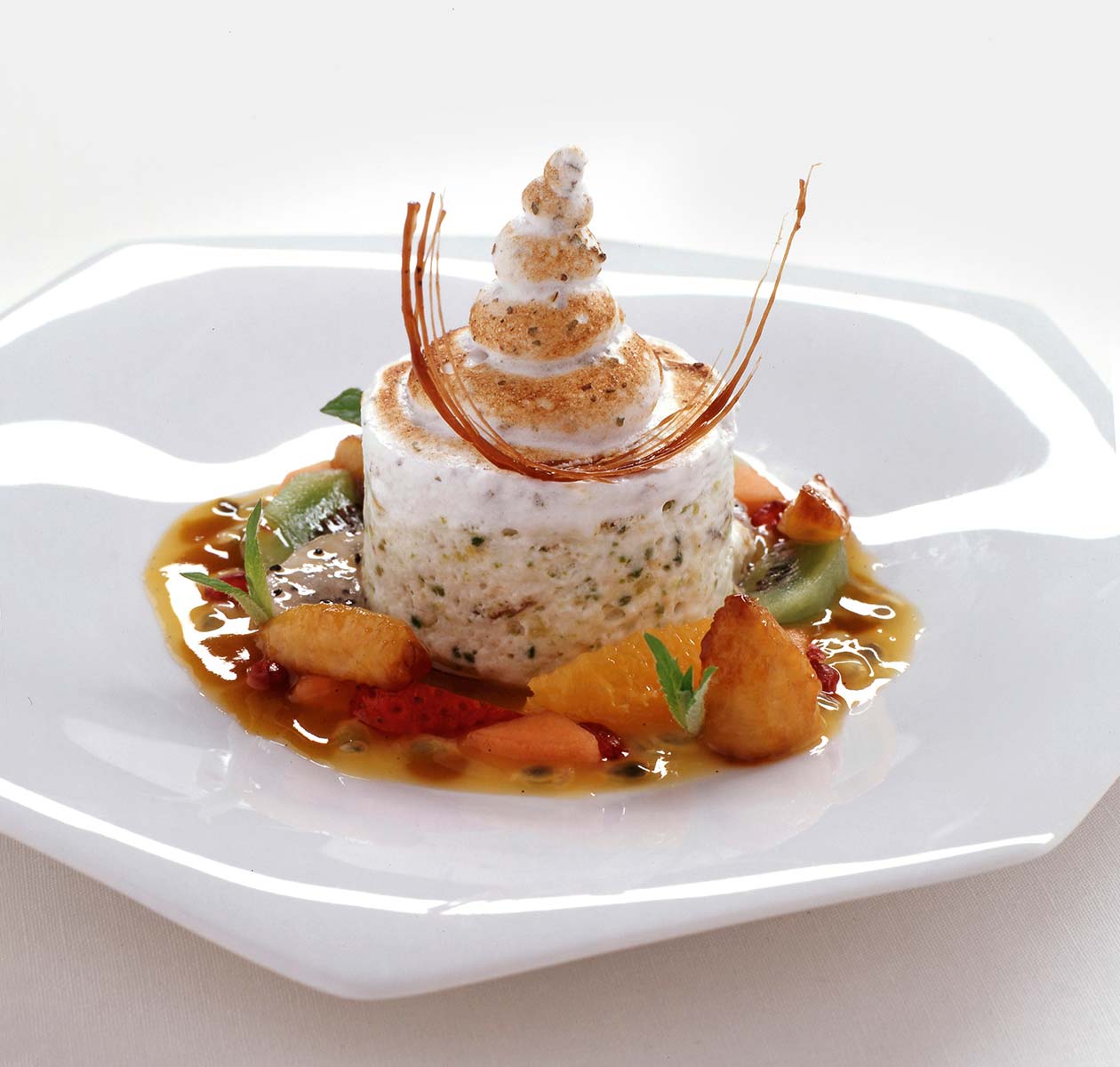 In diesem Bild erkennt man ein Dessert von Schlossberg. Das Dessert ist eine weiße Mousseline, diese wurde in der Mitte des Tellers platziert. Das Mousseline befindet sich in einem Obstsalat mit Soße, bestehend aus Kiwi, Orange, Erdbeere, Melone.