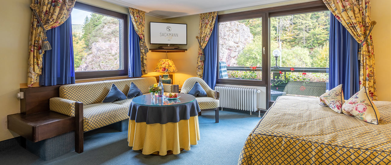 Das Sackmann Doppelzimmer Charme. Im Vordergund ist links angeschnitten das Bett zu erkennen welches Kissen mit Blumenmuster sowie eine gelb blaue Decke besitzt. In der Mitte des Bildes sieht man einen Tisch welcher aus einer Glasplatte und einer gelb blauen Tischdecke besteht. Hinter dem Tisch steht eine Couch sowie ein Sessel, diese besitzen ein gelb blaues Karo und blaue Kissen. Im Hintergrund auf der rechten Seite kann man durch das große Fenster und die Balkontür den Balkon sehen. 